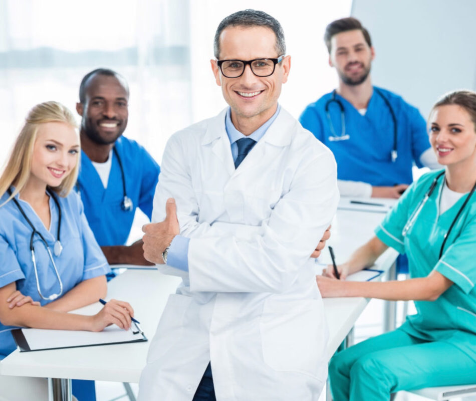 handsome general practitioner with crossed arms with colleagues sitting at table on background